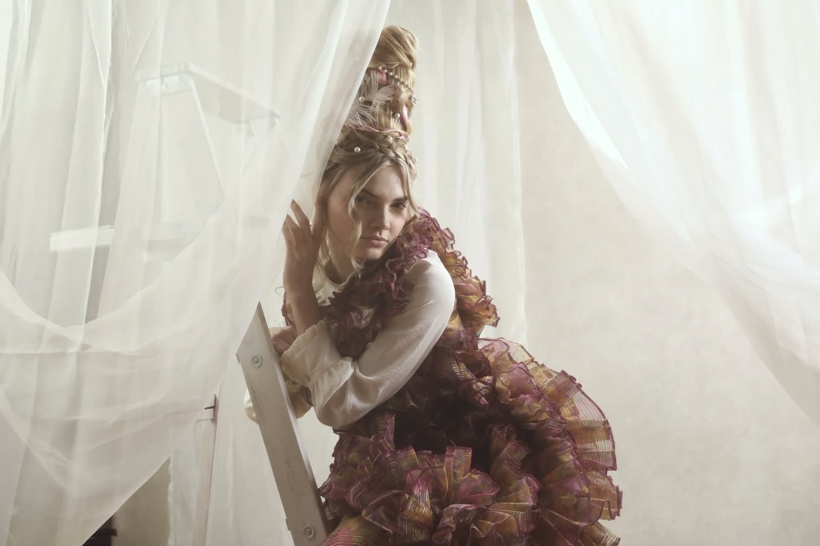 Woman with a tall blonde updo sits among white drapes in a ruffled plum and gold organza top