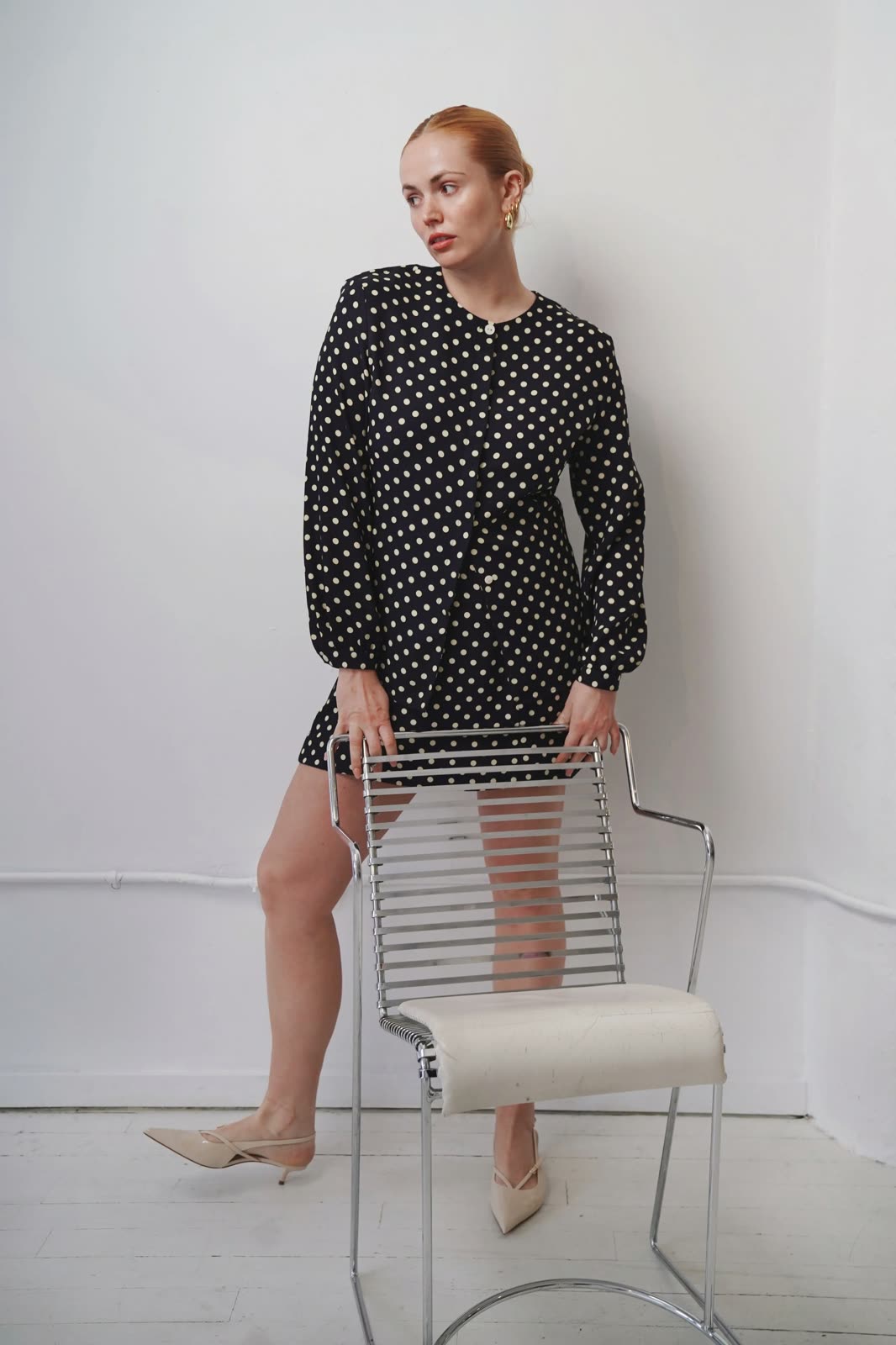 Chloie Costello in a black polka-dot dress and cream heels, one foot on a chrome chair
