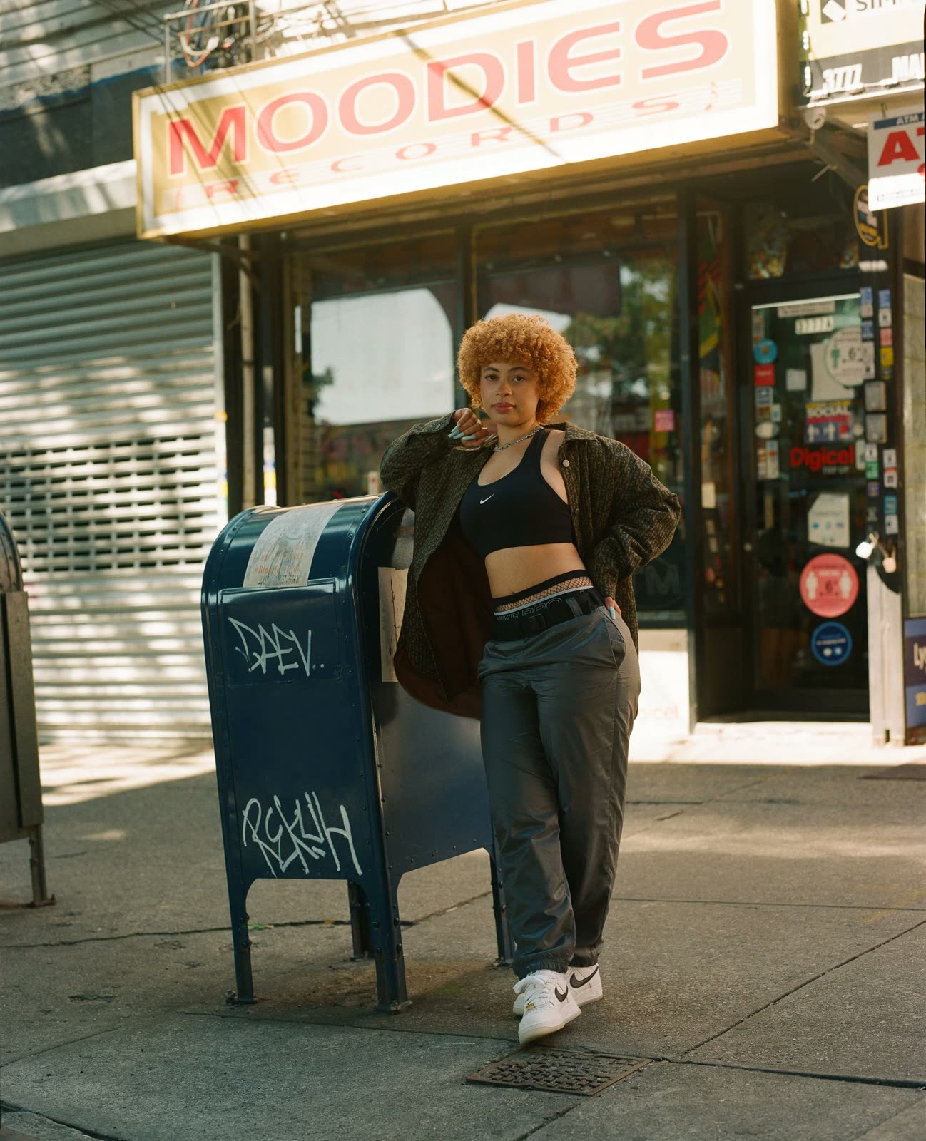 Ice Spice in a black crop top, olive jacket and grey cargo pants by a mailbox outside Moodies Records