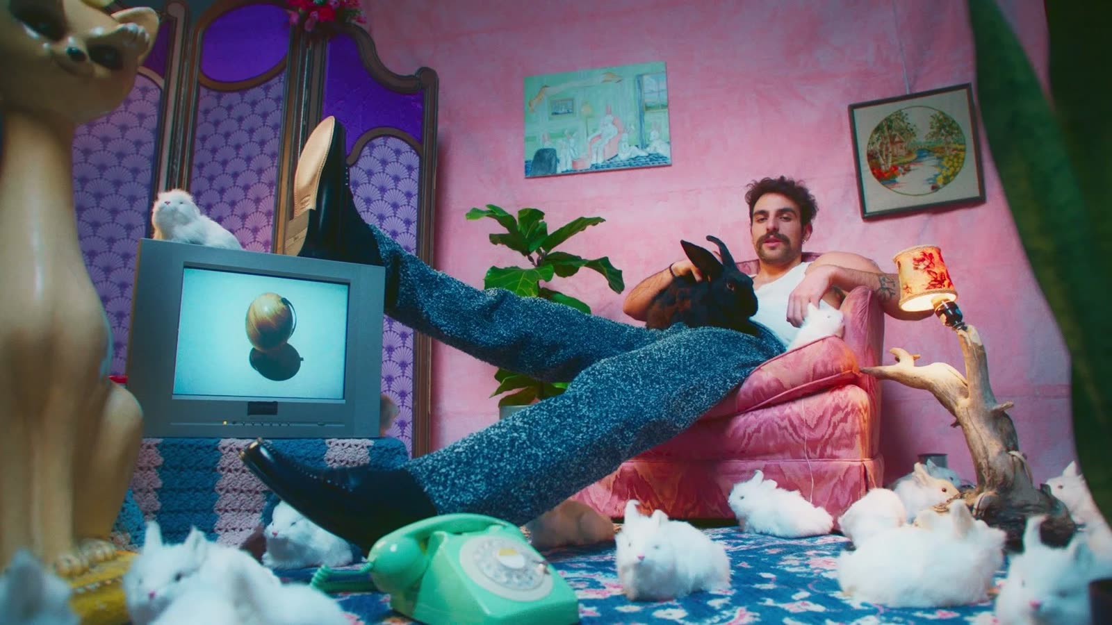 Wide shot of a man in speckled knit trousers in a pink room with white rabbits, Facetime video still