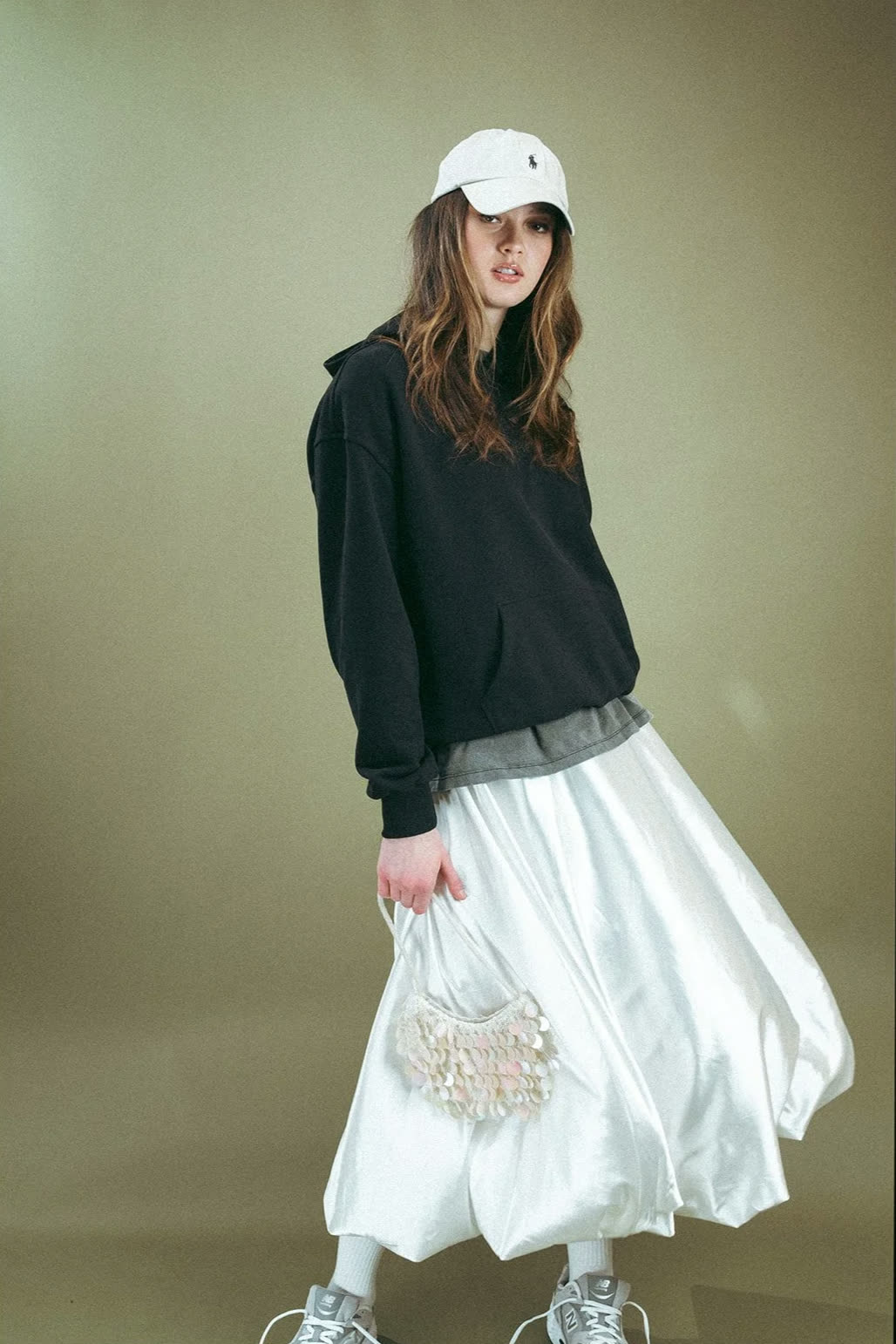 Woman in a white cap, black hoodie and white satin bubble skirt holds a beaded bag